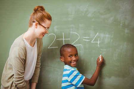 Rear view of teacher assisting little boy to write on blackboard in the classroomの写真素材