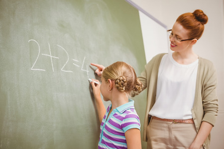 Side view of teacher assisting little girl to write on blackboard in the classroomの写真素材