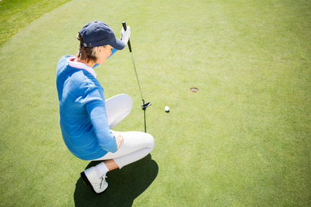 Focused lady golfer kneeling on the putting green on a sunny day at the golf courseの写真素材
