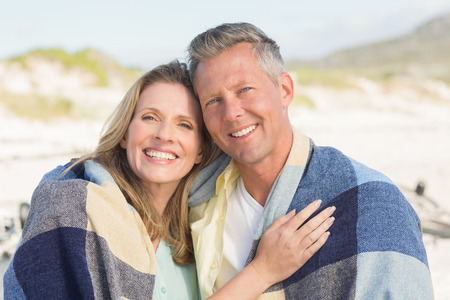 Happy couple wrapped up in blanket at the beachの写真素材