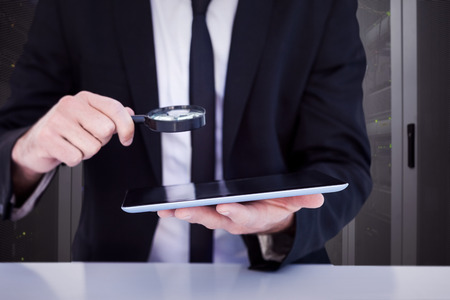 Businessman looking at his tablet through magnifying glass against data center