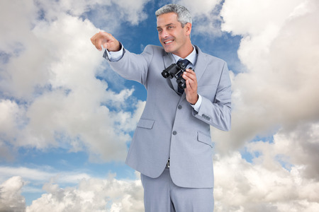 Businessman holding binoculars and pointing out something against blue sky with white cloudsの写真素材