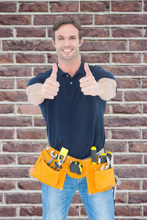 Man wearing tool belt while showing thumbs up sign against red brick wallの写真素材