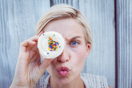 Pretty blonde woman grimacing with cupcake on wooden backgroundの写真素材