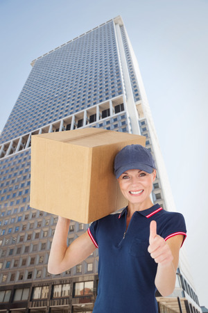 Happy delivery woman holding cardboard box showing thumbs up against skyscraperの写真素材