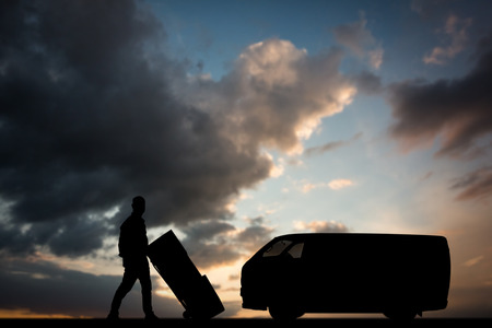 Happy delivery man pushing trolley of boxes against blue and orange sky with cloudsの写真素材