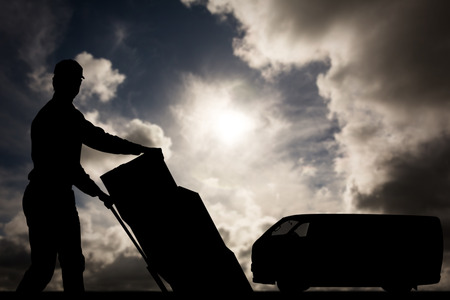 Happy delivery man pushing trolley of cardboard boxes against dark sky with white cloudsの写真素材