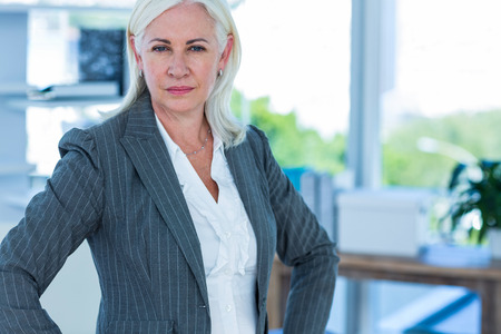 Serious businesswoman looking at camera with hands on hips in officeの写真素材