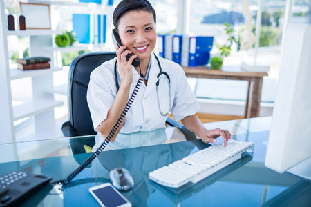 Smiling doctor having phone call and using her computer in medical officeの写真素材