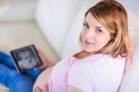 Pregnant woman looking at camera with hand on belly and ultrasound scans in the living roomの写真素材
