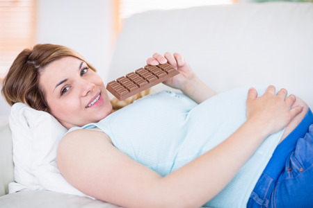 Pregnant woman looking at camera and eating chocolate at home in the living roomの写真素材