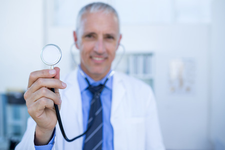 Happy doctor smiling at camera and showing his stethoscope in medical officeの写真素材