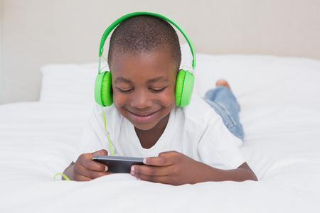 Pretty little boy using smartphone and listening music in bed at home in bedroomの写真素材