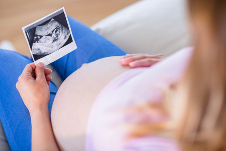 Pregnant woman looking at ultrasound scans and touching her belly in the living roomの写真素材