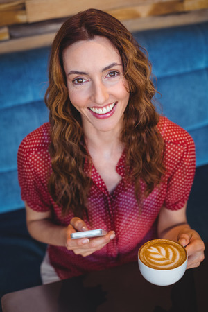 Woman using her mobile phone and holding cup of coffee at the coffee shopの写真素材
