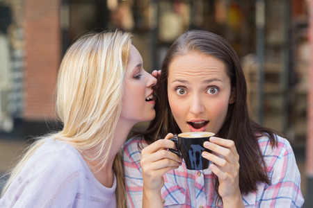 Blonde woman telling secret to her friend while drinking coffee outside at a cafe