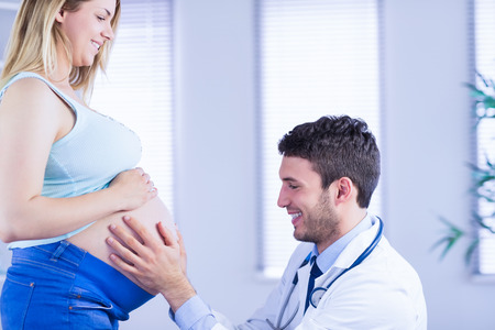 Smiling doctor checking stomach of standing pregnant patient in medical officeの写真素材