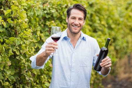 Young happy man smiling at camera and holding a glass of wine in the grape fieldsの写真素材