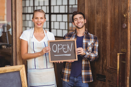 Portrait of smiling waitress and man holding chalkboard with open sign at coffee shop