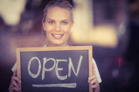Portrait of smiling blonde waitress showing chalkboard with open sign at coffee shopの写真素材