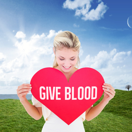 Attractive young blonde showing red heart against green field under blue skyの写真素材