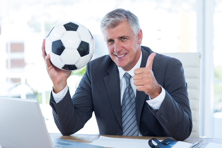 Smiling businessman holding soccer ball with thumbs up in his officeの写真素材