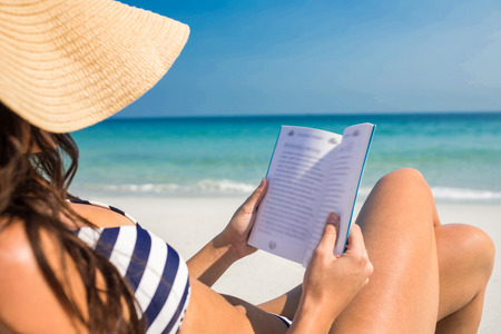 Pretty brunette reading on deck chair at the beach on a sunny dayの写真素材