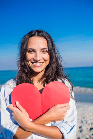 Smiling woman holding heart card at the beach on a sunny dayの写真素材