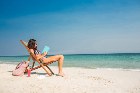 Pretty brunette reading a book on deck chair at the beachの写真素材