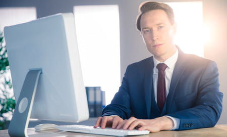 Confident businessman typing on computer and looking at camera in his officeの写真素材