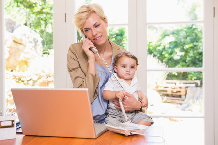 Pretty blonde woman with his son phoning and using laptop in the officeの写真素材