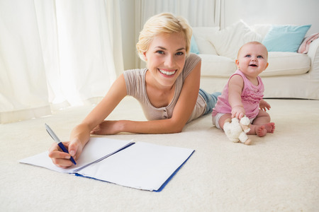 Happy blonde mother with her baby girl writting on a copybook at home in the living roomの写真素材