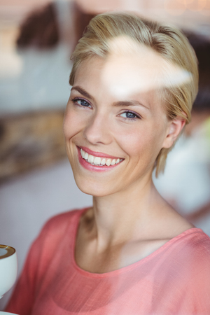 Happy blonde woman smiling at camera and holding a cup of coffee at the coffee shopの写真素材