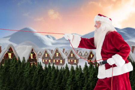 Santa pulls something with a rope against cute village in the snowの写真素材