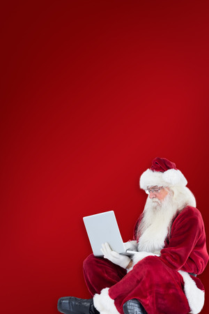 Santa sits and uses a laptop against red backgroundの写真素材