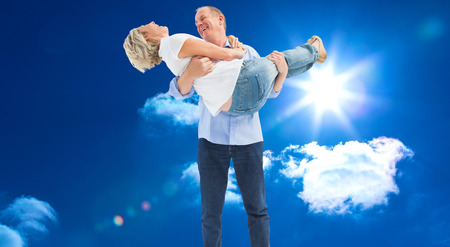 Mature man carrying his laughing partner against bright blue sky with cloudsの写真素材