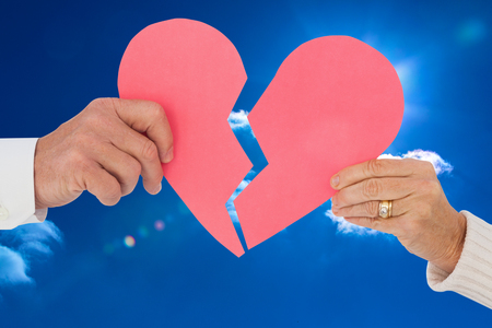 Couple holding a broken paper heart against bright blue sky with cloudsの写真素材