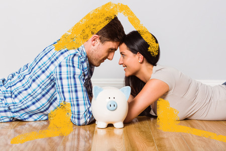 Young couple lying on floor smiling with piggy bank against house outlineの写真素材