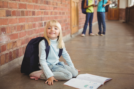 Portrait of cute pupil kneeling over notepad at corridor in school