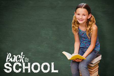 Cute little girl reading book in library against green chalkboard
