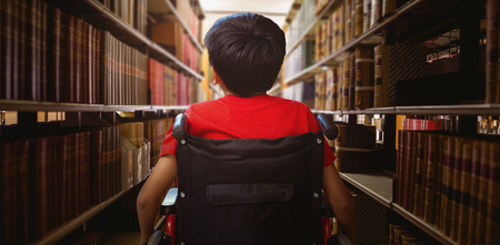 Rear view of boy sitting in wheelchair against close up of a bookshelfの写真素材