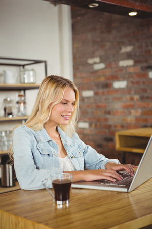 Smiling blonde having coffee and using laptop at coffee shopの写真素材