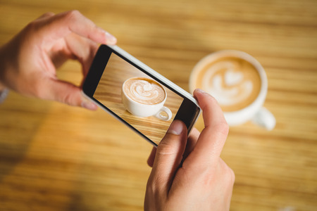 Man photographing his cappuccino with coffee art at coffee shopの写真素材