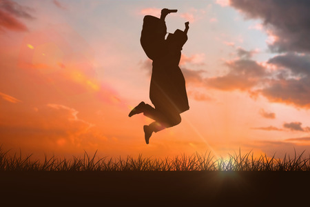 Happy male student in graduate robe jumping against orange and blue sky with cloudsの写真素材