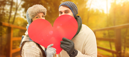 Attractive young couple in warm clothes holding red heart against autumn sceneの写真素材