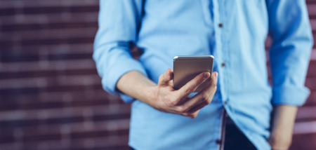 Midsection of man holding cellphone against brick wallの写真素材