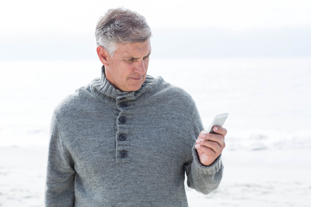 Smiling man standing and on his phone at the beachの写真素材