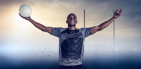 Confident athlete with arms raised holding rugby ball against goals postsの写真素材