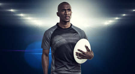 Thoughtful athlete holding rugby ball against spotlightsの写真素材