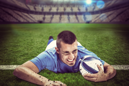 Confident rugby player lying in front with ball against large football stadium with lightsの写真素材